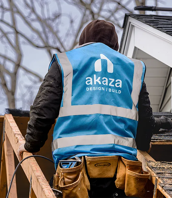 A construction professional from Akaza Builders wearing a blue safety vest and tool belt stands on wooden roof rafters during a residential project, representing a premier design-build general contractor in Central MA.
